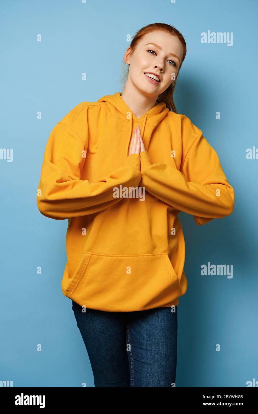 Red-haired girl in a yellow sweatshirt stands on a blue background holding her folded palms in front of her Stock Photo
