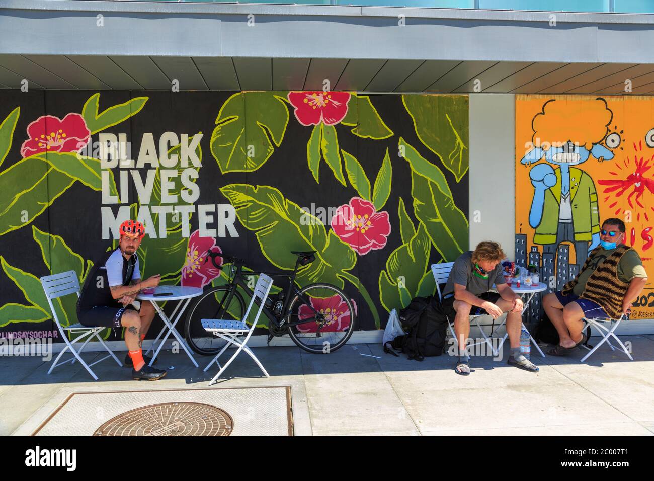 People sitting outdoors at a cafe in front of painted murals protesting the murder of George Floyd on plywood boards covering storefront windows on Lincoln Blvd, Santa Monica, Los Angeles,  California Stock Photo