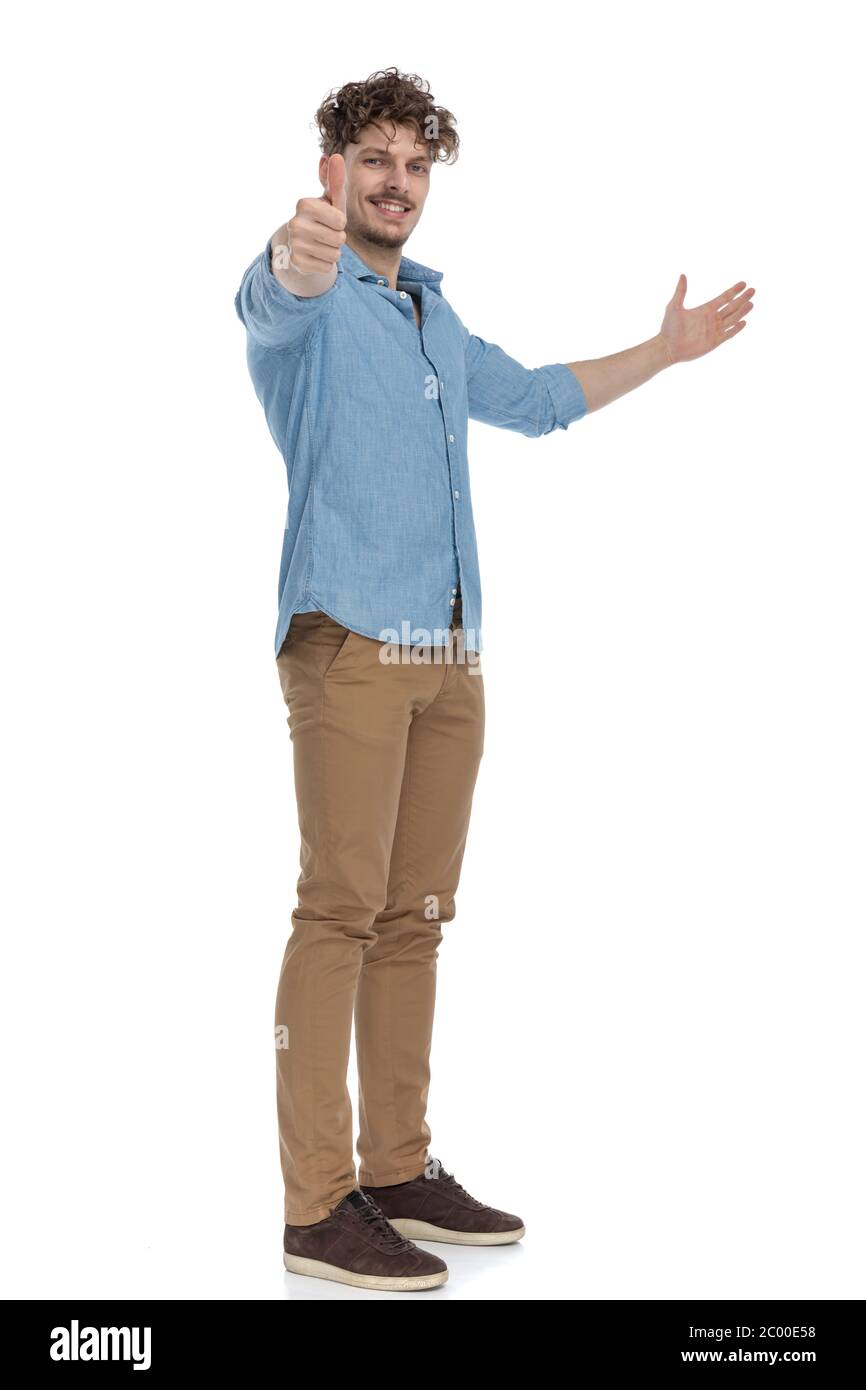 casual young man smiling, inviting to side and making thumbs up sign ...