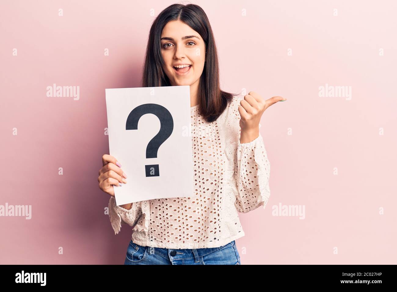 Young beautiful girl holding question mark pointing thumb up to the ...
