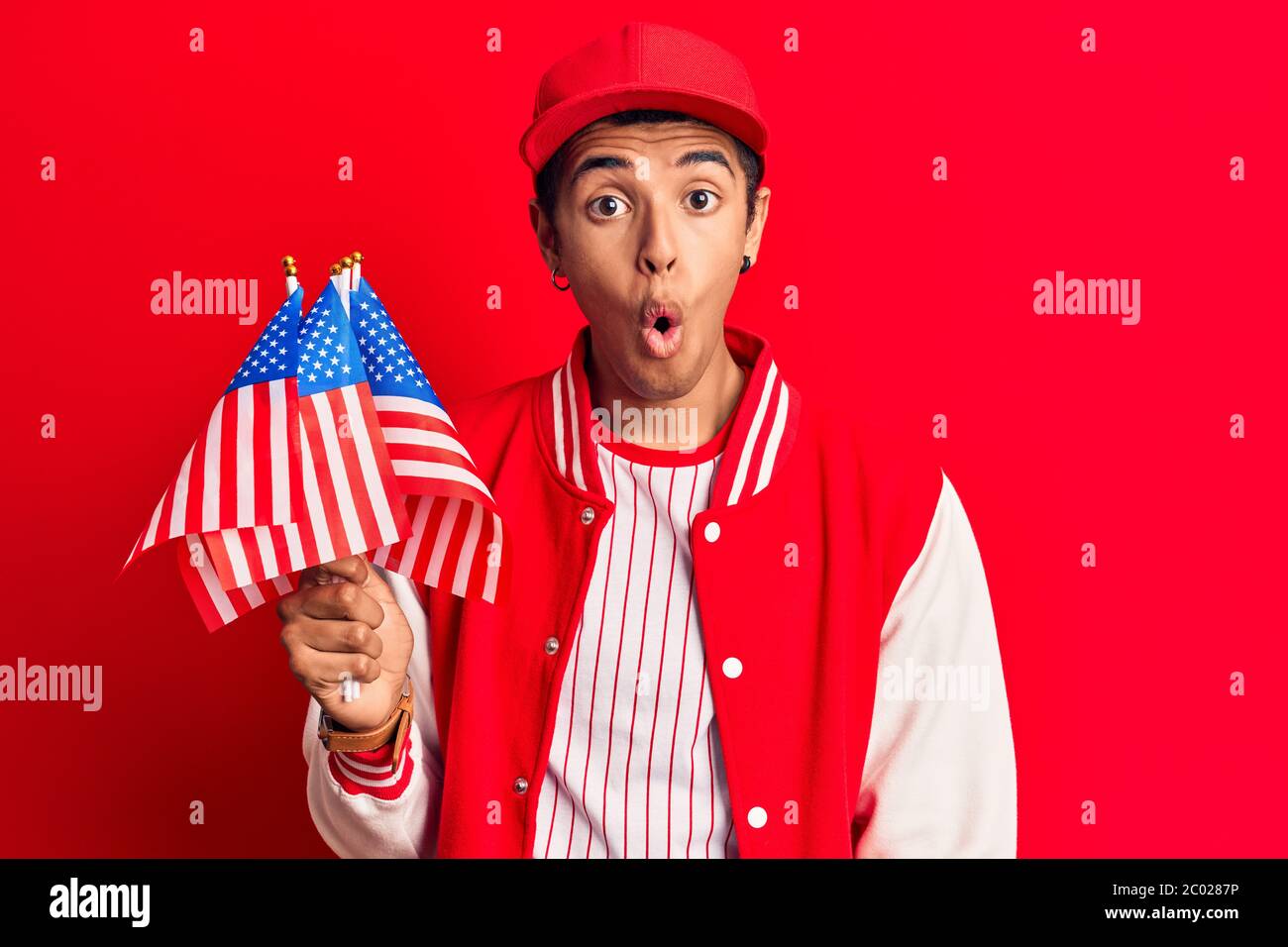 Young african amercian man wearing baseball uniform holding america flags scared and amazed with open mouth for surprise, disbelief face Stock Photo