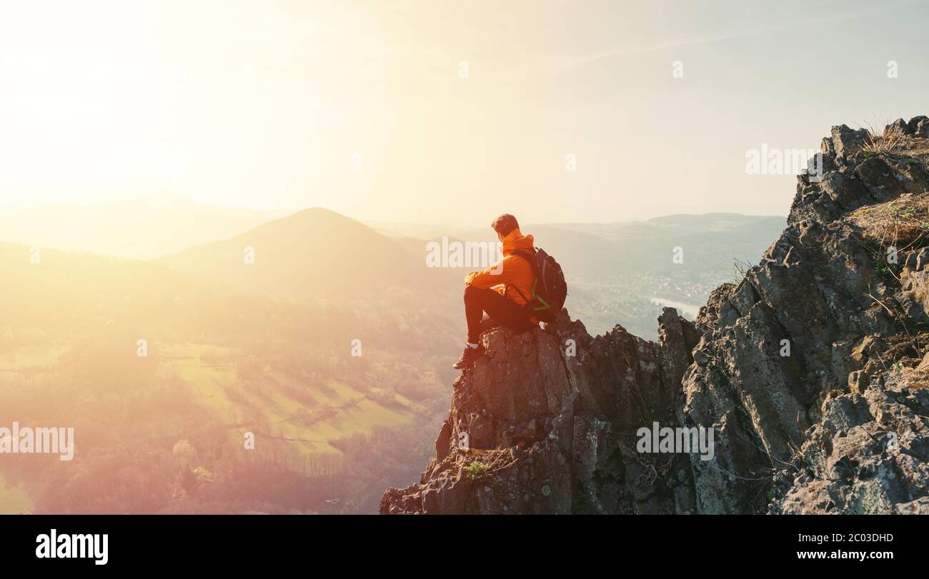 Travel man tourist alone on the edge cliff mountains and looking on the valley. Silhouette of the person on the high rock at sunset. Hiking adventure Stock Photo