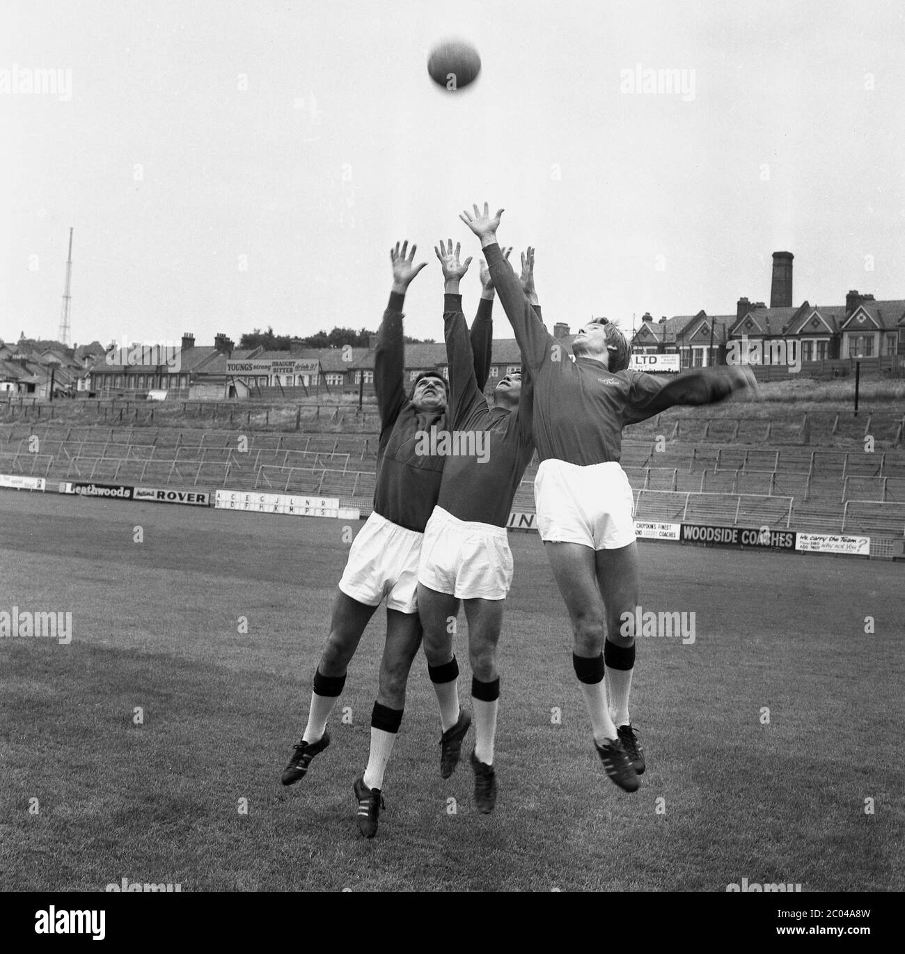 1960s, historical, three goalkeepers leaping for the ball as they train together on the pitch at Crystal Palace FC, Croydon, South London, England, UK. The large banked terraces with metal barriers where football supporters stood and watched a match in this era can be seen in the background. Stock Photo