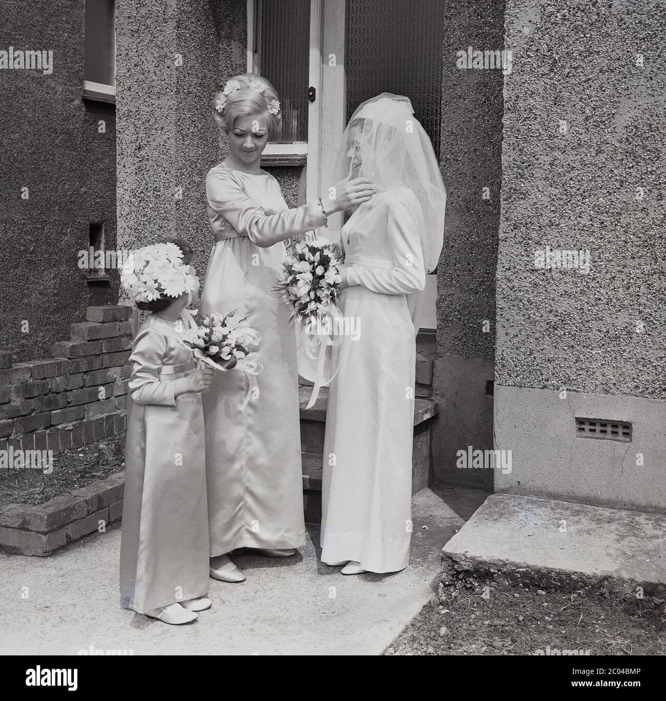 1960s, historical, last minute preparations from her chief bridesmaid for the young bride in her wedding dress and veil standing outside the front door in the porch of a pebble-dash suburban house, South London, England, UK. A little flower girl stands with them holding her bouquet. Stock Photo