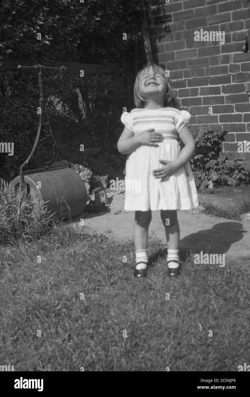 1940s, historical, 'What a lovely day'......a happy little girl wearing a summer dress standing on a lawn in a back garden looking up to sky with her eyes closed taking in the lovely sunshine, Sheffield, England, UK. A traditional heavy lawn roller with metal handle is seen in the picture. Stock Photo