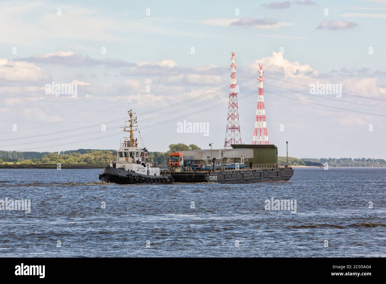 Tug boat TAUCHER O. WULF pushing  deck cargo pontoon T.O.W.II with extra wide heavy load truck on board on Elbe river near Stade Stock Photo
