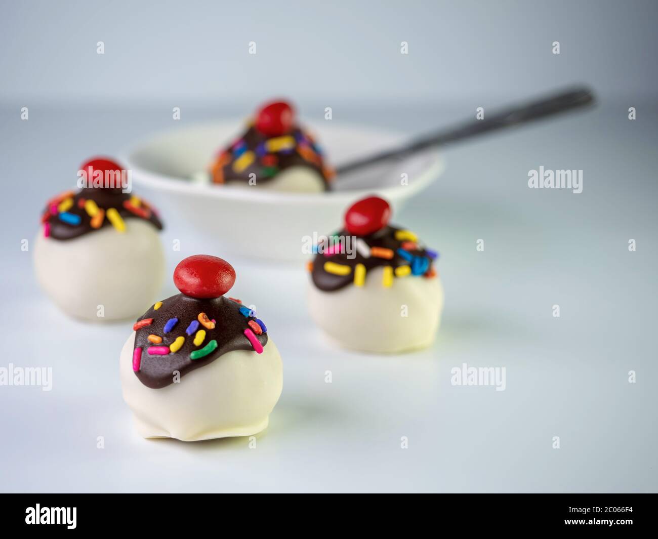 Banana split cake balls with white coating, chocolate drizzle covered in sprinkles and a cherry on top with a white bowl and spoon. Stock Photo