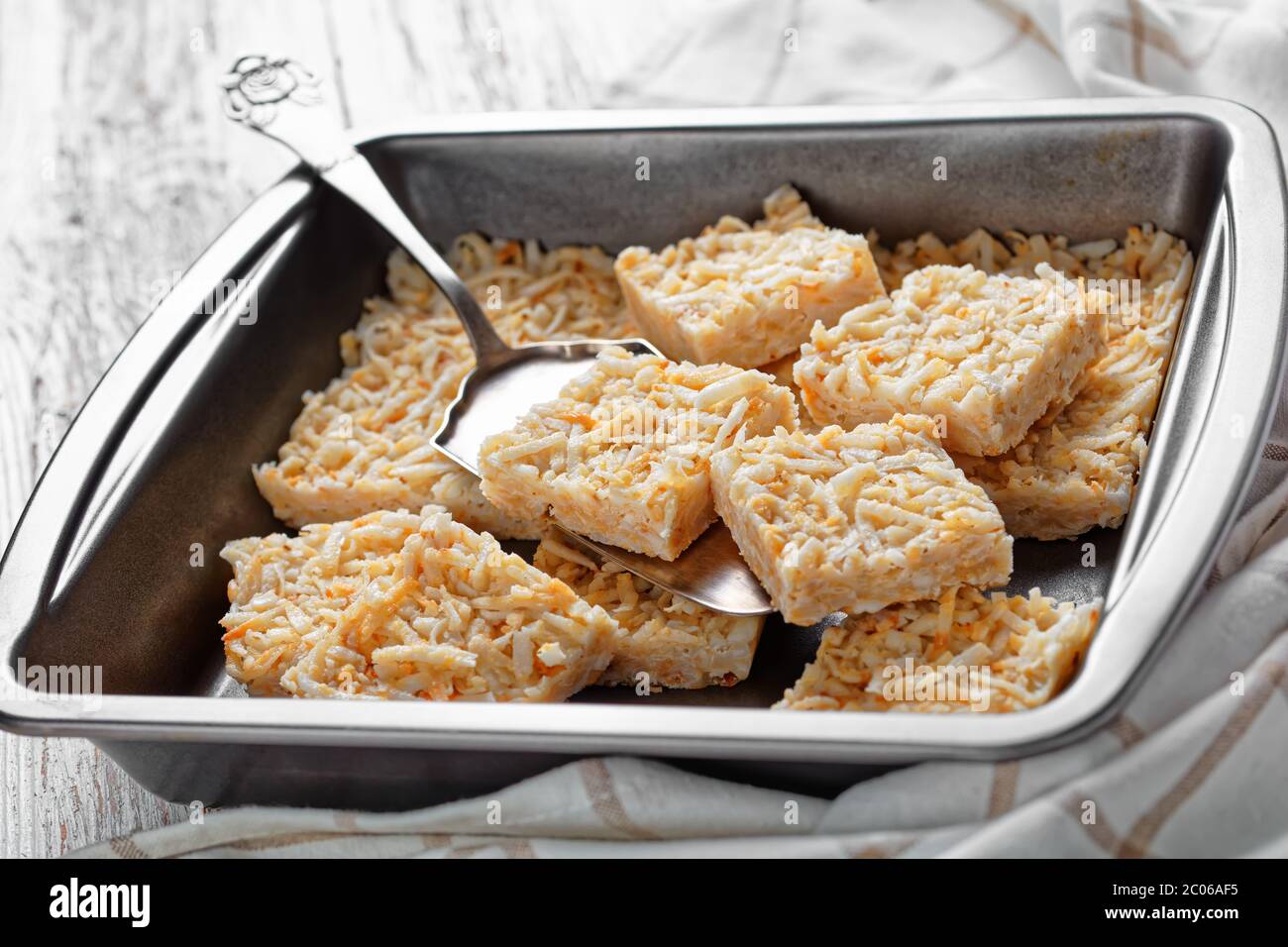 close-up of no-bake coconut crack bars in a baking dish and one on a cake shovel on a rustic wooden table Stock Photo