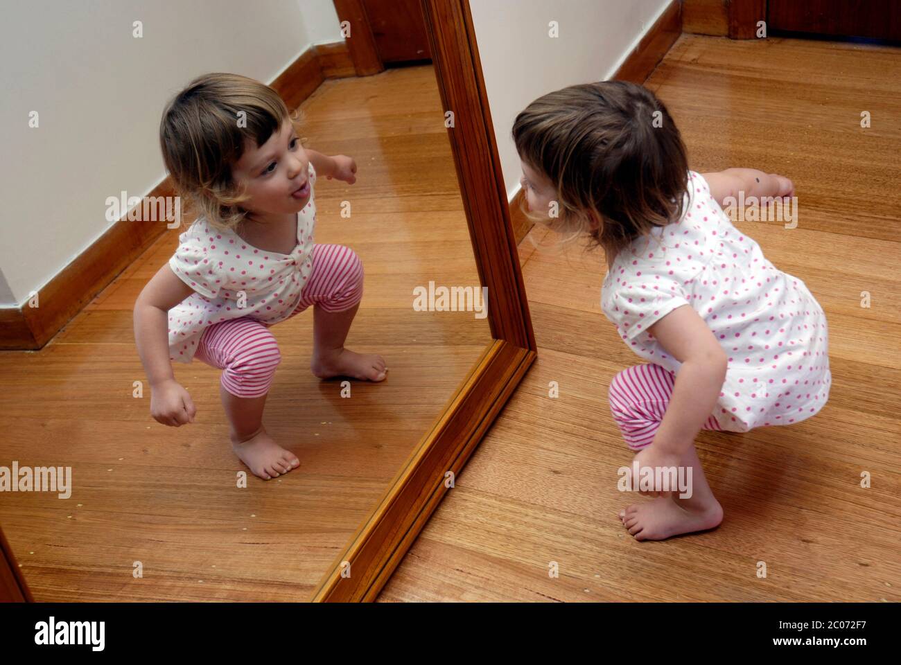 A young girl, two and a half years old, playing in front of a large mirror. Stock Photo