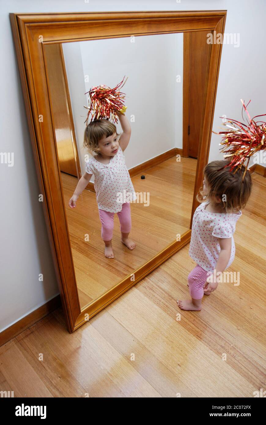 A young girl, two and a half years old, playing in front of a large mirror. Stock Photo