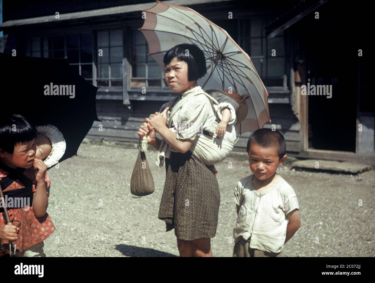 [ 1950s Japan - Girls carrying Babies ] — Young girls with umbrellas carrying babies, ca. 1950 (Showa 25).  They are most likely carrying their siblings, but they could also be komori.  Komori were young girls from poor families who were hired by the year to aid middle and upper class mothers in the care of their infant. In addition to carrying the baby around, the komori would also assist a little with the household work.  Komori received food, lodging, clothing, and occasionally some money.  20th century vintage slide film. Stock Photo