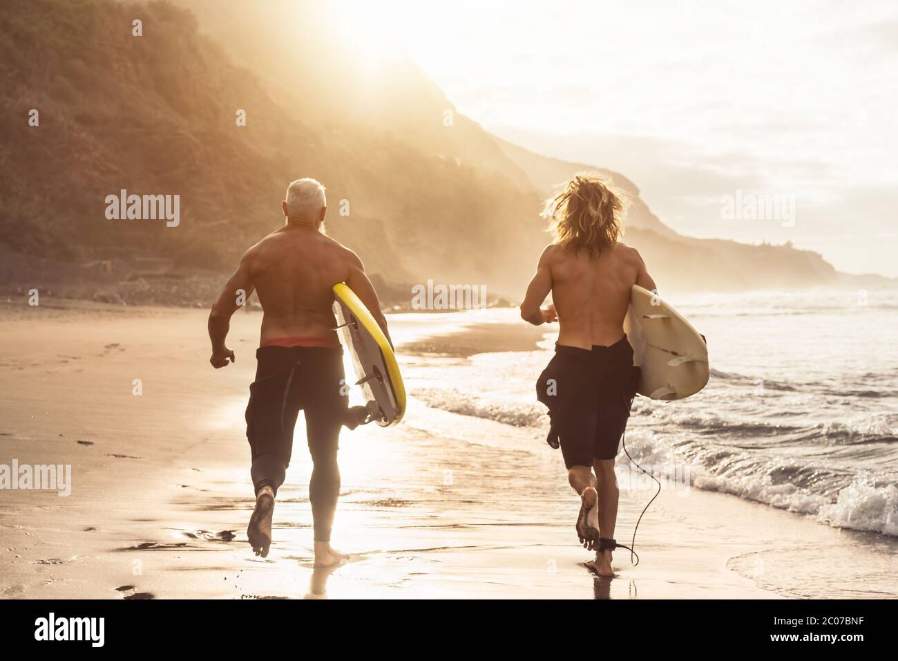 Happy fit friends having fun surfing on sunset time - Surfers father and son running out the ocean Stock Photo