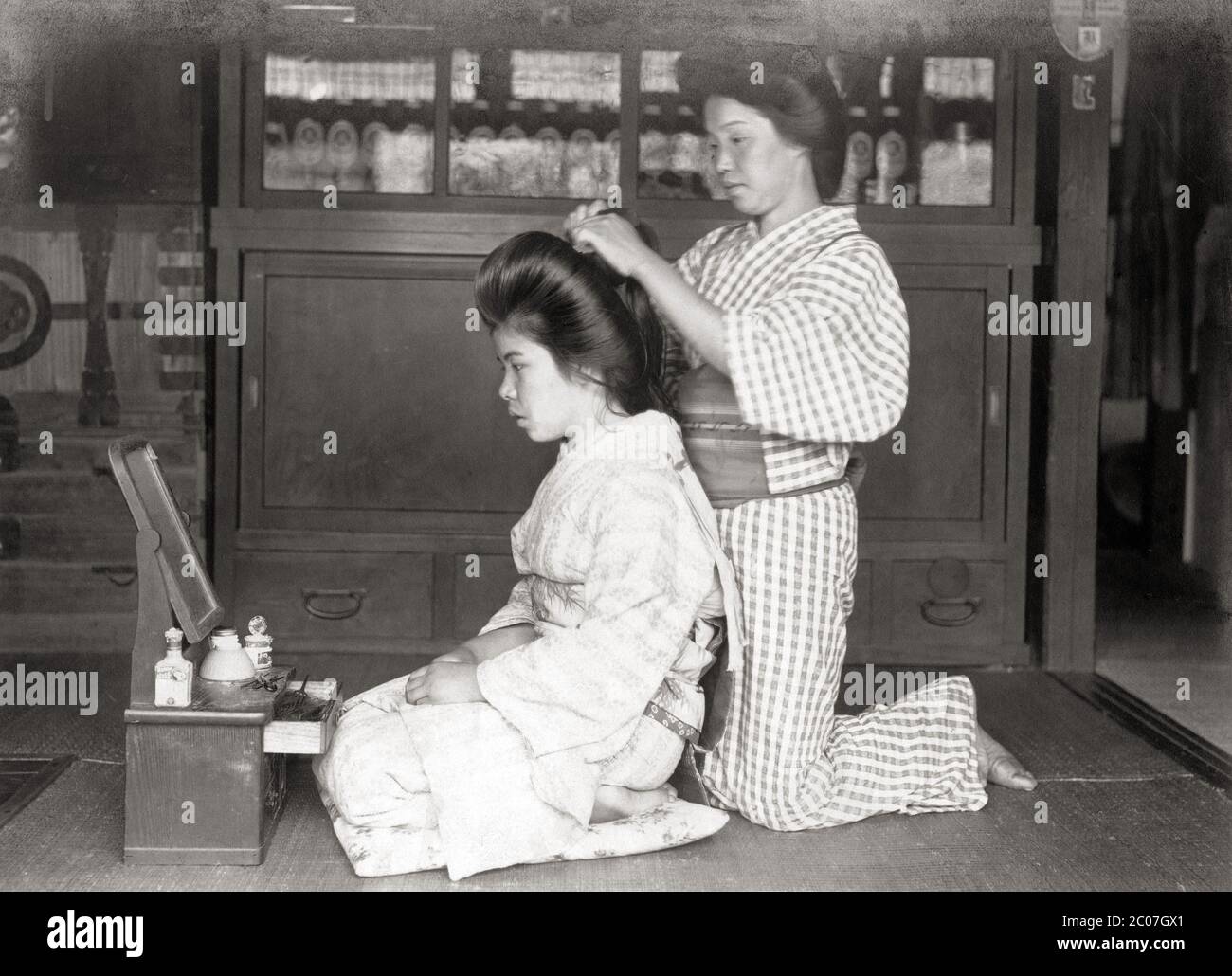 [ 1910s Japan - Hairdresser at Work ] — A woman dressed in a kimono and facing a mirror has her hair done by a hairdresser.  20th century vintage gelatin silver print. Stock Photo