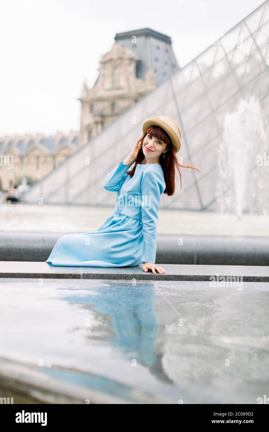 PARIS, FRANCE - September 17, 2019: Young happy woman in blue dress, sitting near the fountain and posing for a photo with the glass pyramid Stock Photo