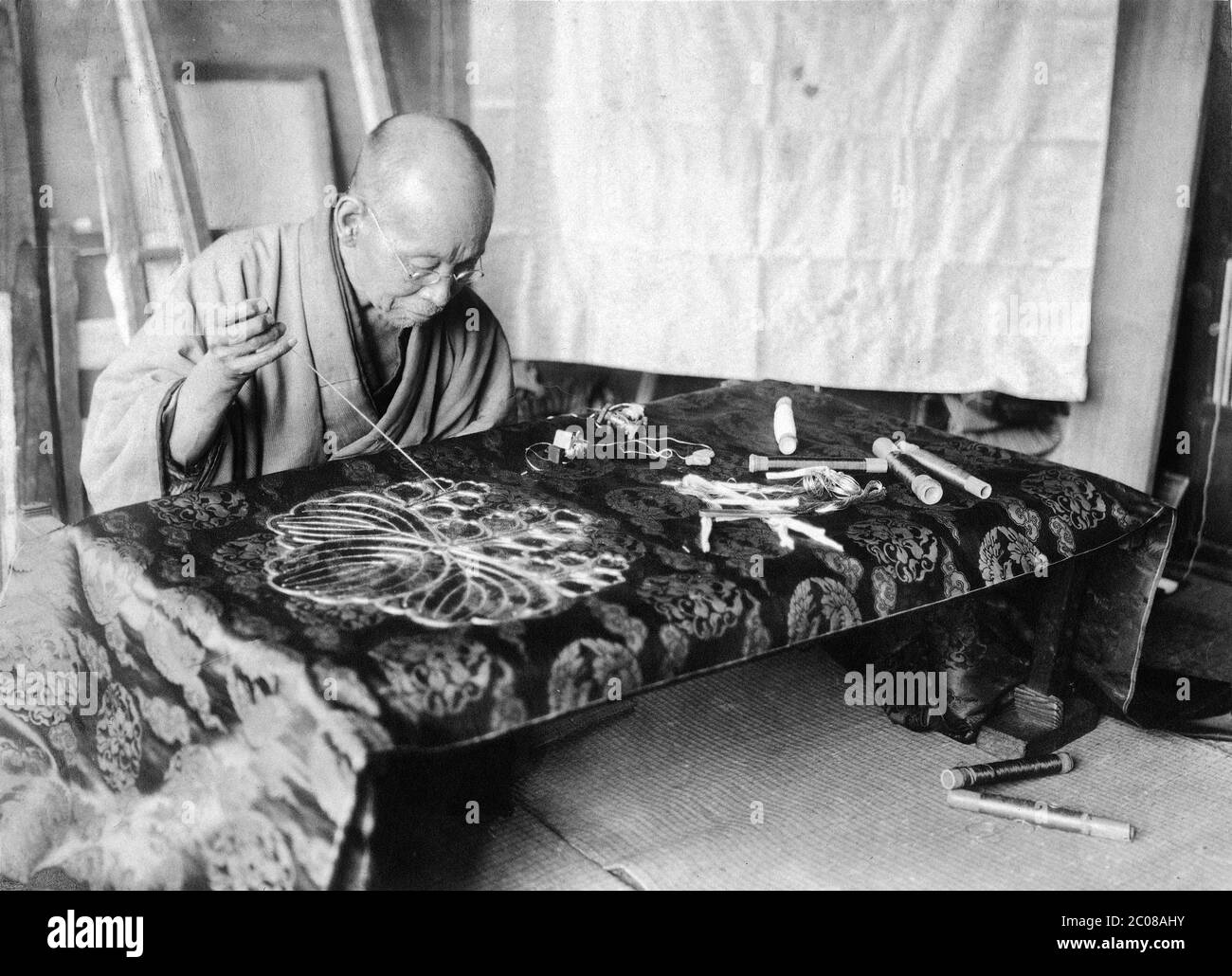 [ 1910s Japan - Artisan Embroidering Obi ] — A craftsman in traditional clothing is embroidering a silk obi, a long, broad sash tied around the waist of a kimono.  20th century vintage gelatin silver print. Stock Photo