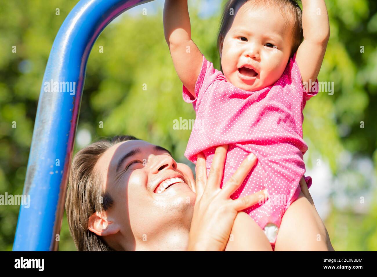 Happy dad helping his kid hang on the monkey bars at the park on a sunny day. Stock Photo