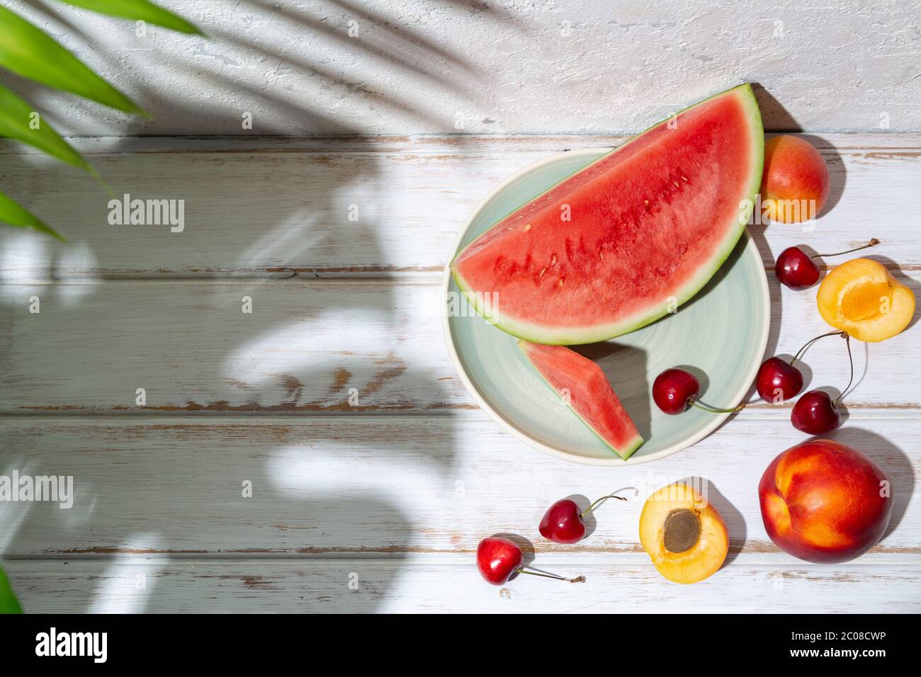 Summer fruit composition with tropical leaves shadows on a white wooden table and concrete wall. Copy space Stock Photo