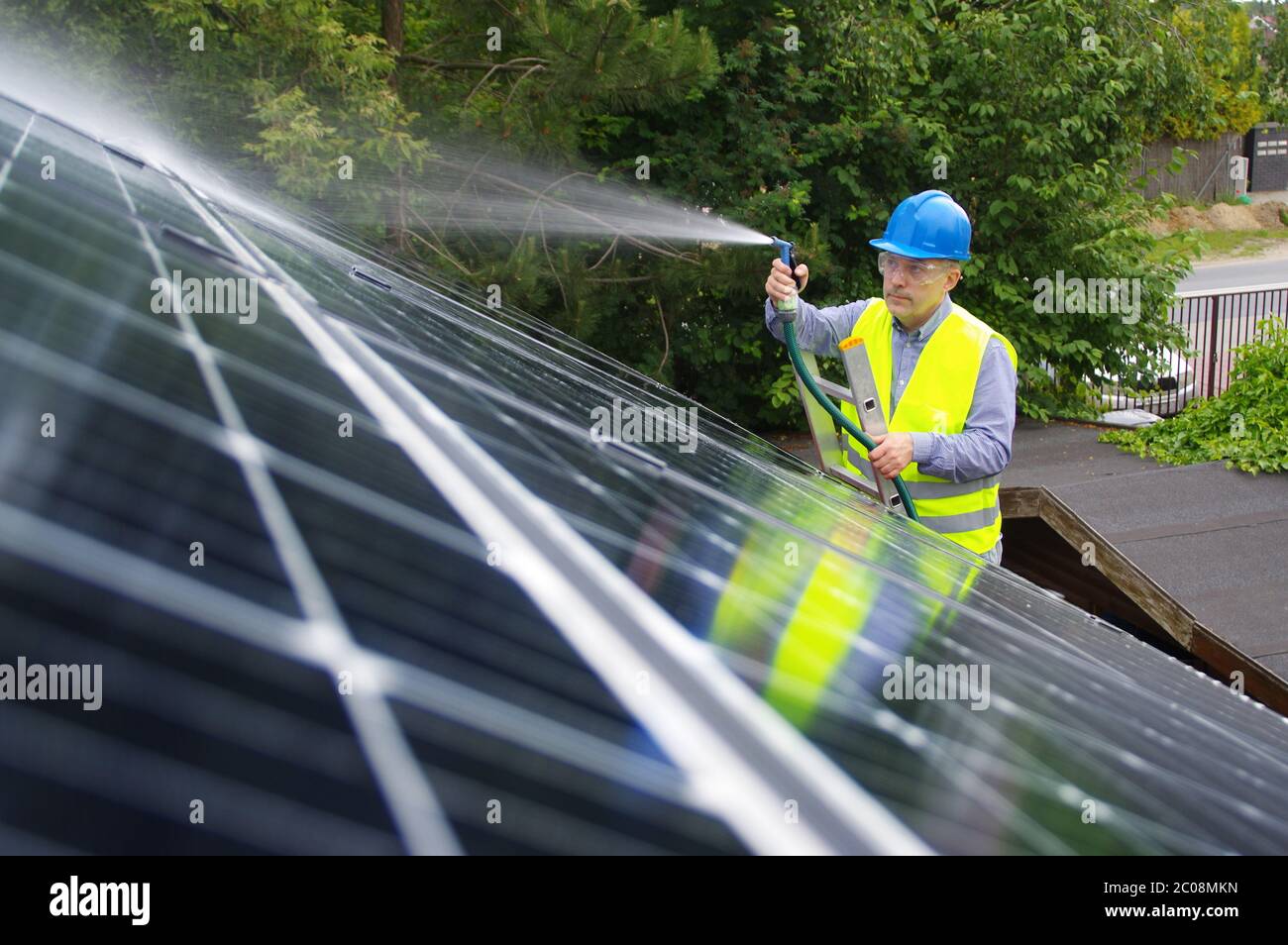 Man cleans solar panel on the roof. Maintenance of renewable energy in a private home. Worker washes the solar installation with water. Stock Photo