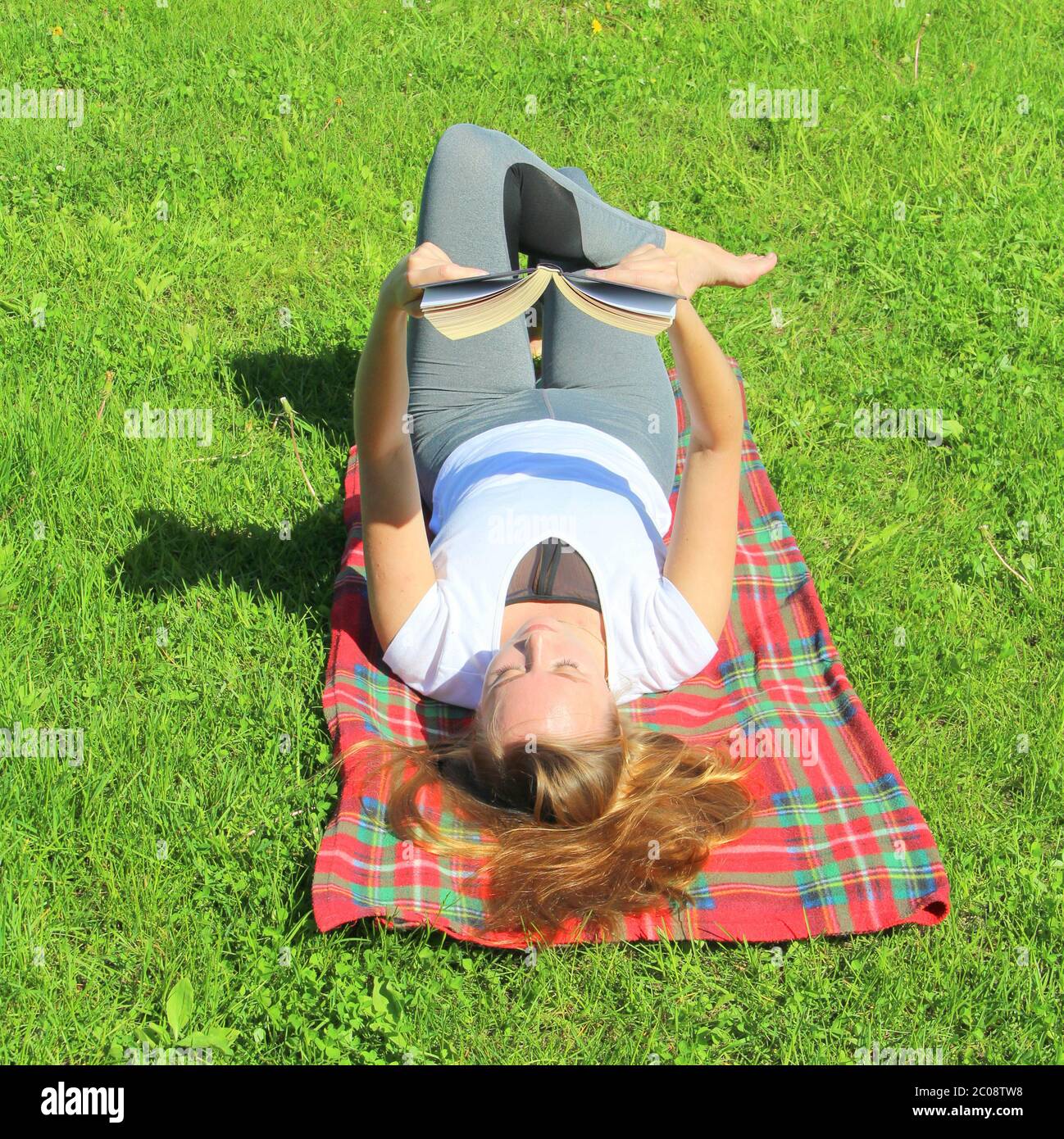A beautiful young white girl in a white T-shirt and with long hair lies on a red plaid, on green grass, on the lawn and read book. Stock Photo