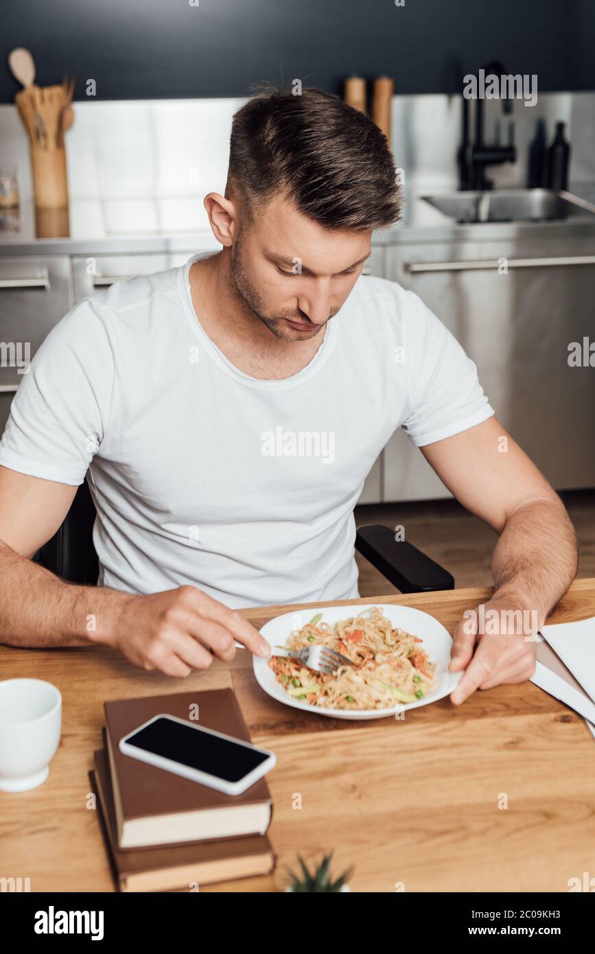 Selective focus of man eating noodles near smartphone on books on kitchen table Stock Photo