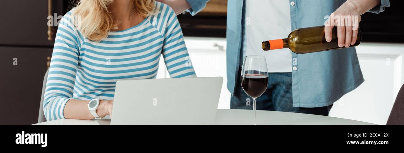 Panoramic shot of man pouring wine near woman using laptop in kitchen Stock Photo