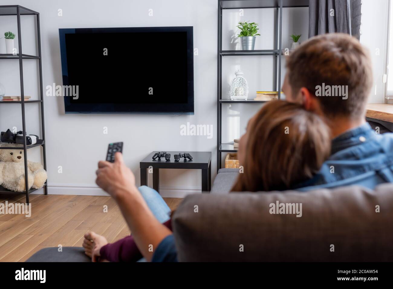selective focus of man holding remote controller and looking at flat panel tv with blank screen near woman in living room Stock Photo