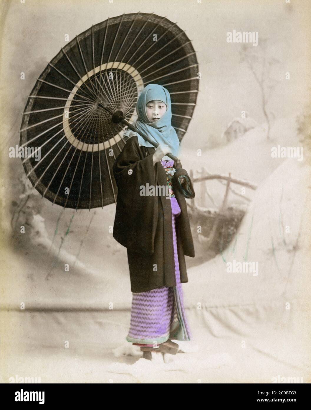[ 1890s Japan - Woman in Winter Outfit ] — A young Japanese woman in a thick winter kimono has an okosozukin (御高祖頭巾) wrapped around her head to protect herself against the winter cold.  She is holding a paper parasol.  19th century vintage albumen photograph. Stock Photo