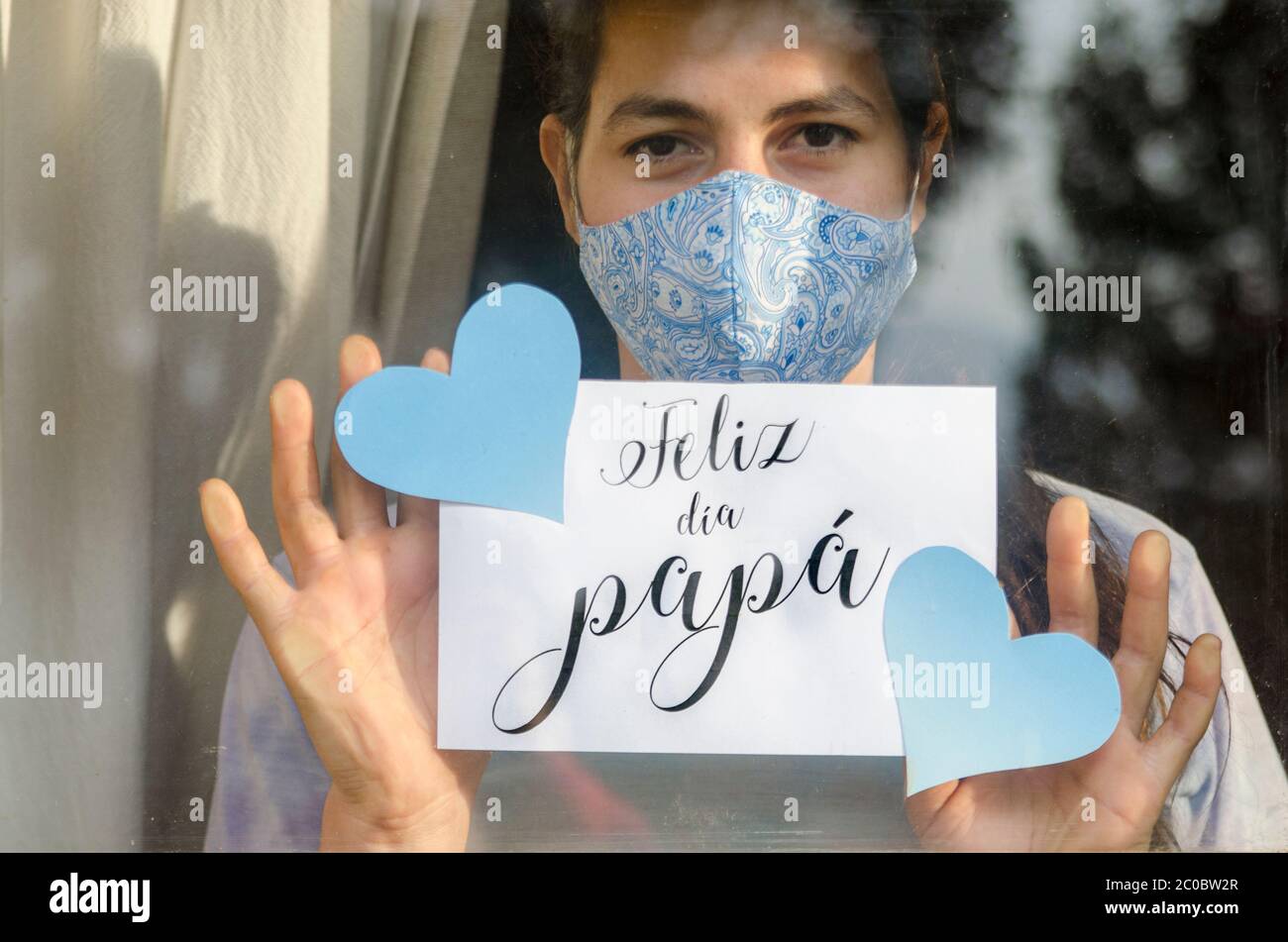 Young girl wearing a facemask at home holding a sheet of paper through the window that says in spanish: 'Happy day, dad!' Stock Photo