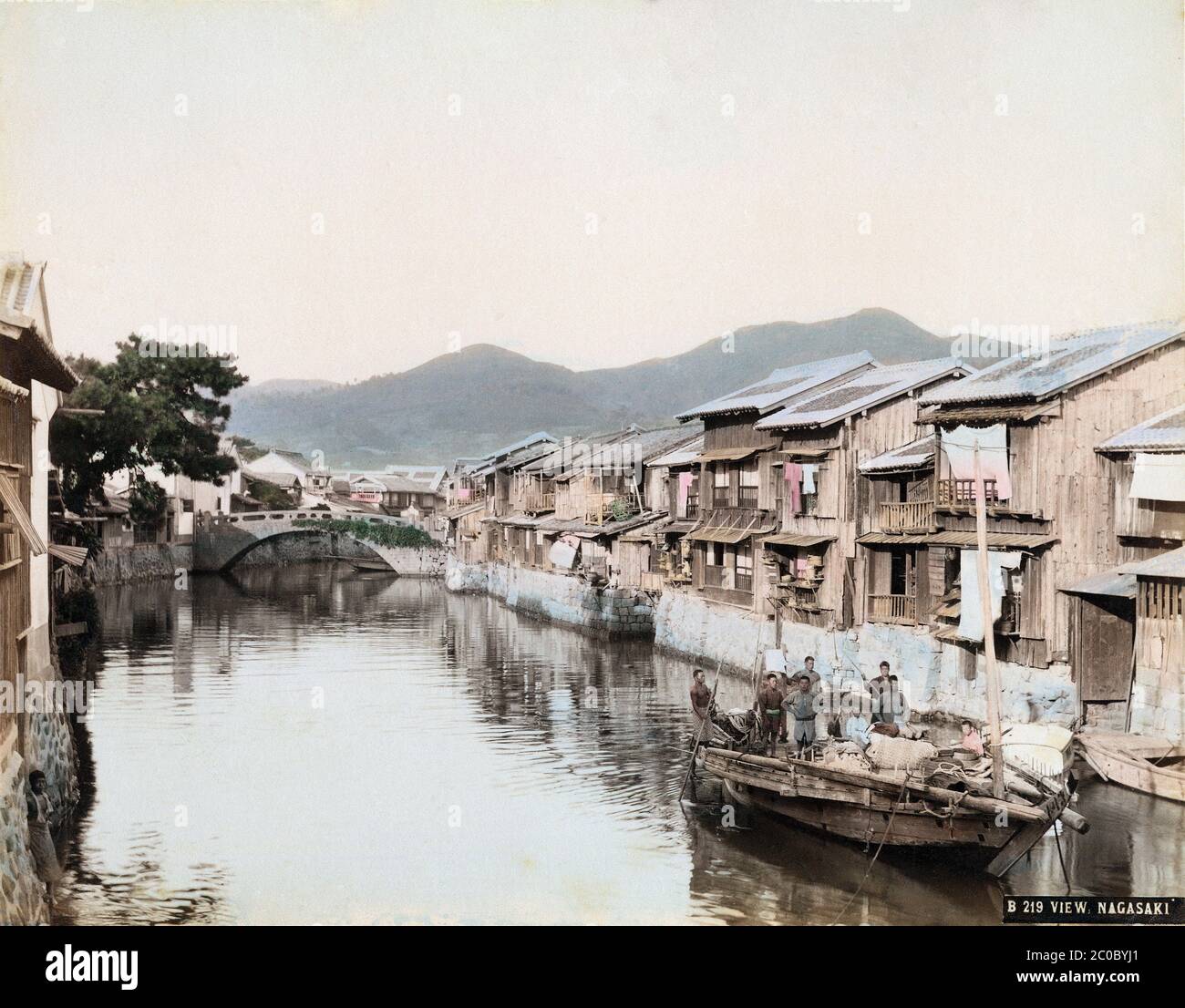 [ 1890s Japan - Boat on Nagasaki River ] — Wooden boat on the Nakashima River (中島川) in Nagasaki.   In the back, the Yorozubashi Bridge (万橋) is visible. Originally built in 1678 (Enpo 6), it was replaced in 1801 (Kansei 13). In 1915 (Taisho 4), the stone bridge on this image was replaced with a concrete bridge with iron girders.  The area on the right is now the streetcar line between Nigiwaibashi Bridge (賑橋) and Nishihamano-machi (西浜町).  19th century vintage albumen photograph. Stock Photo