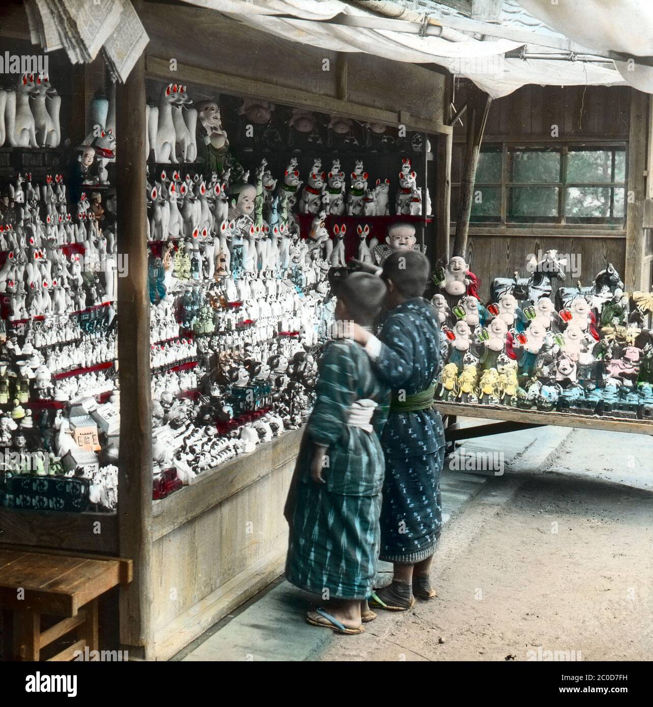 [ 1900s Japan - Japanese Porcelain Shop ] — Two boys look at a display of porcelain dolls and figures at a souvenir shop at Fushimi Inari Taisha, the head shrine of Inari, located in Fushimi-ku, Kyoto.   Most of them are foxes, which are prominent symbols of Inari Okami (稲荷大神), one of the principal kami (deity,  divinity, spirit) of Shinto.  20th century vintage glass slide. Stock Photo