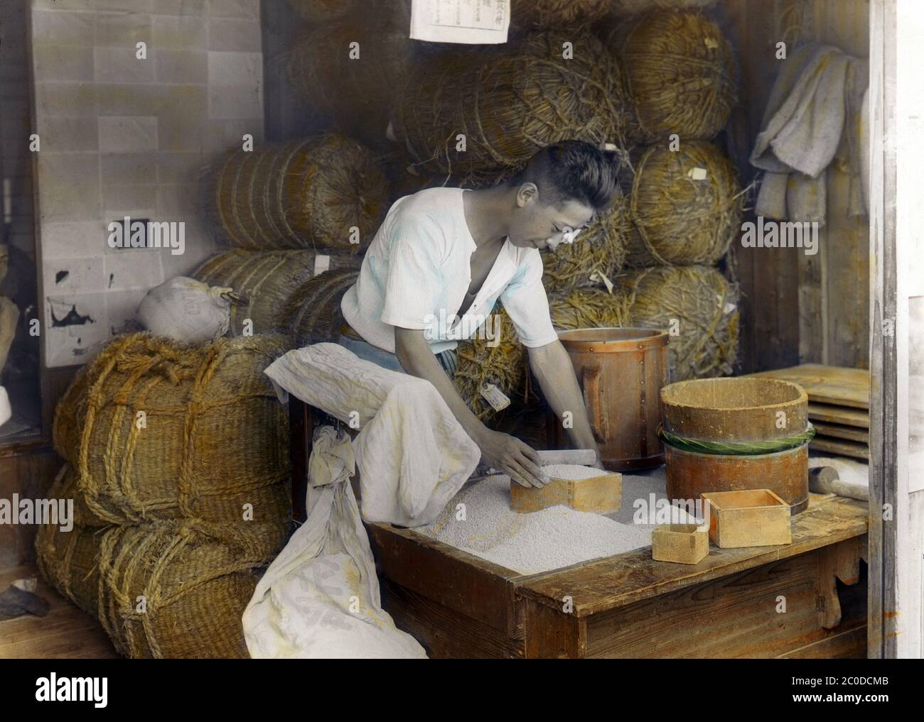 [ 1900s Japan - Japanese Rice Shop ] — A Japanese man measures rice at a rice shop with a square wooden box known as masu (枡 or 升). There are different sized mass on the table.  Behind him, many tawara (俵, straw rice bags) can be seen.  20th century vintage glass slide. Stock Photo