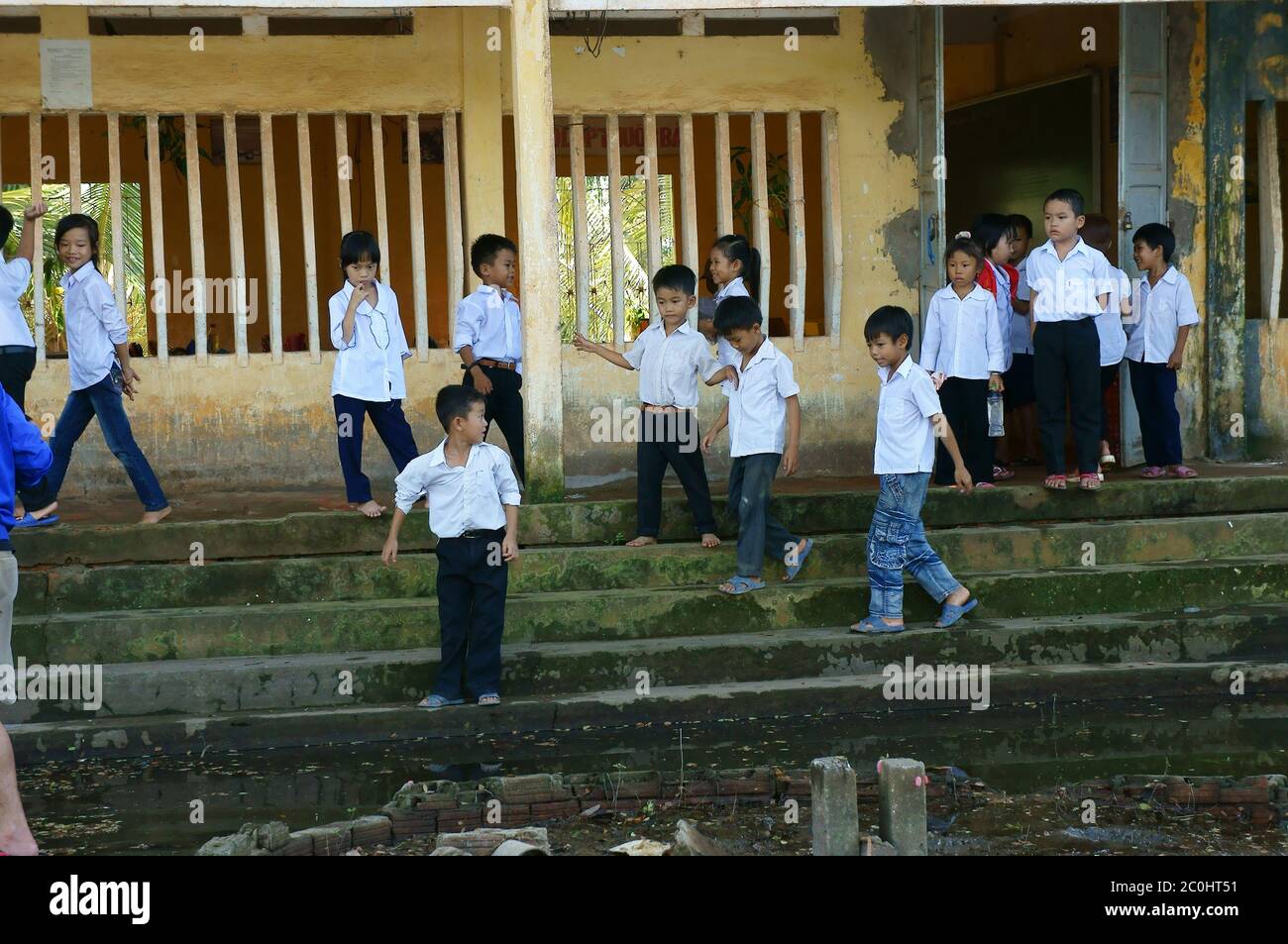 Vietnamese little boy pupils wear uniform: white t shirt, blue trousers standing with fun together on flooded school yard front of poor building Stock Photo