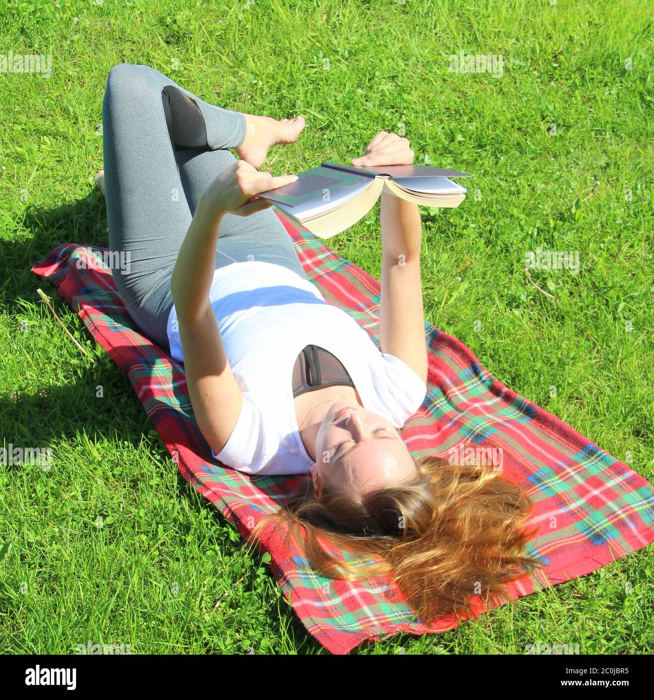 A beautiful young white girl in a white T-shirt and with long hair lies on a red plaid, on green grass, on the lawn and read book. Stock Photo