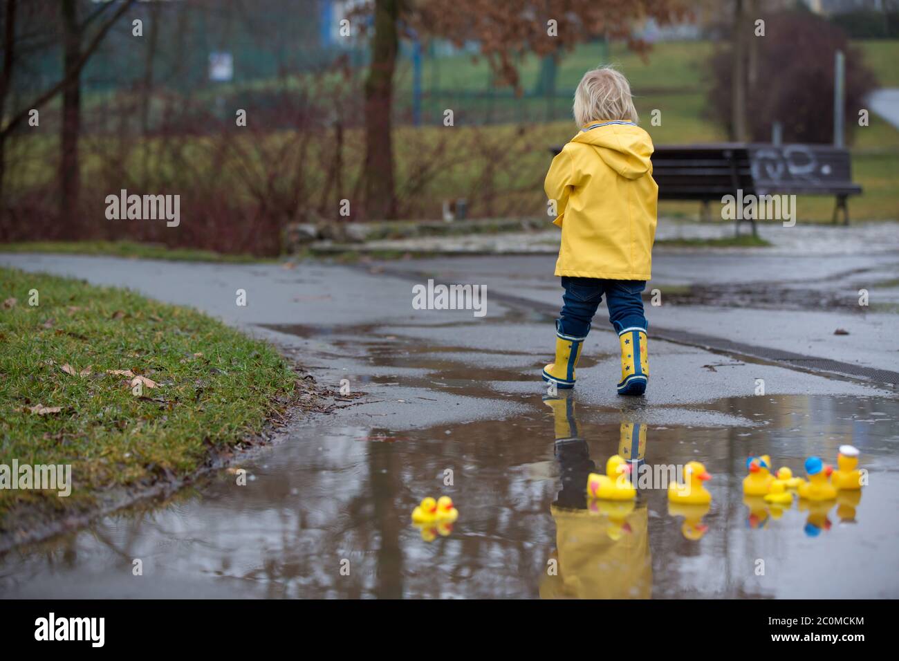 Beautiful funny blonde toddler boy with rubber ducks and colorful umbrella, jumping in puddles and playing in the rain, wintertime Stock Photo