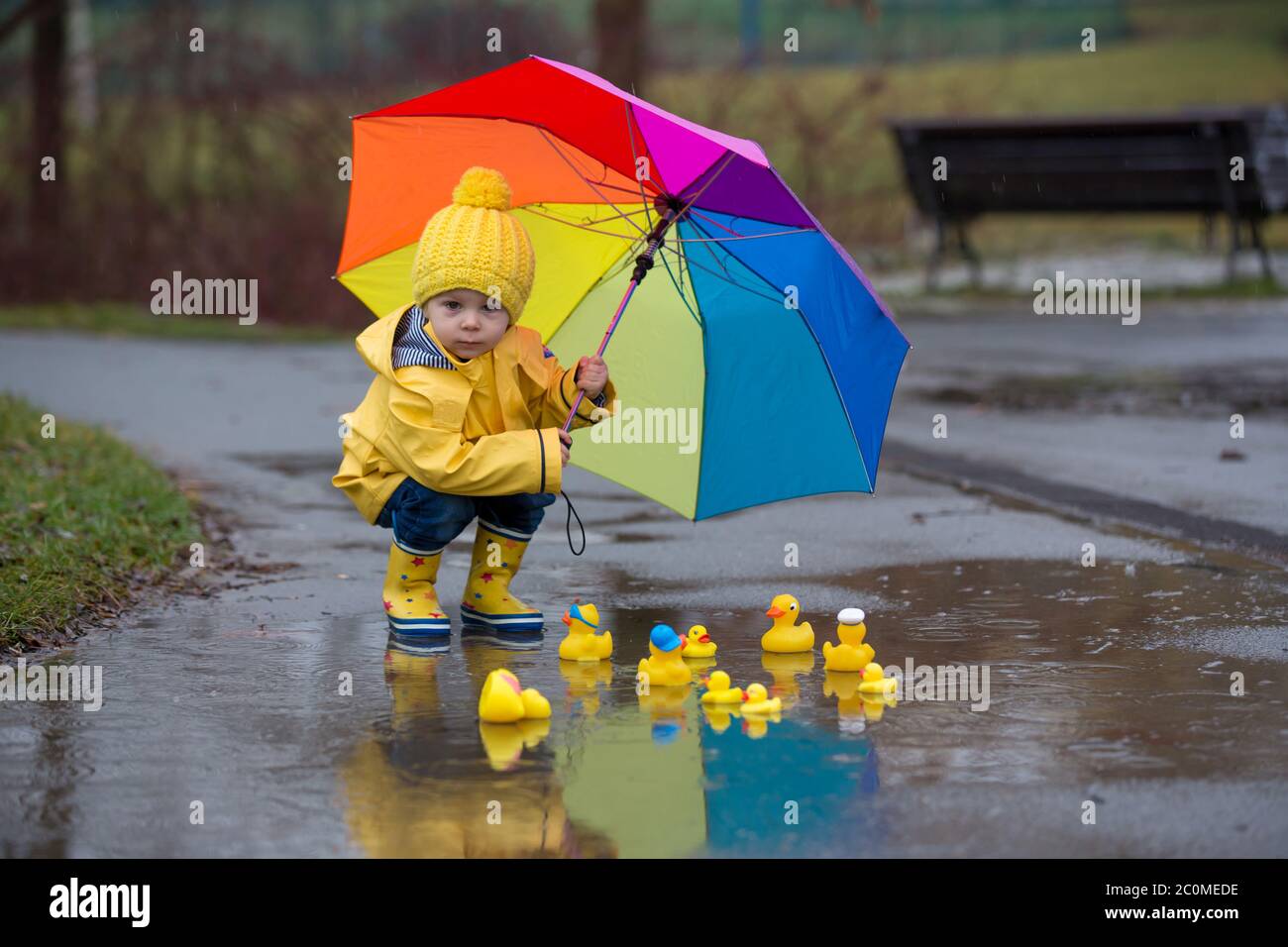 Beautiful funny blonde toddler boy with rubber ducks and colorful umbrella, jumping in puddles and playing in the rain, wintertime Stock Photo