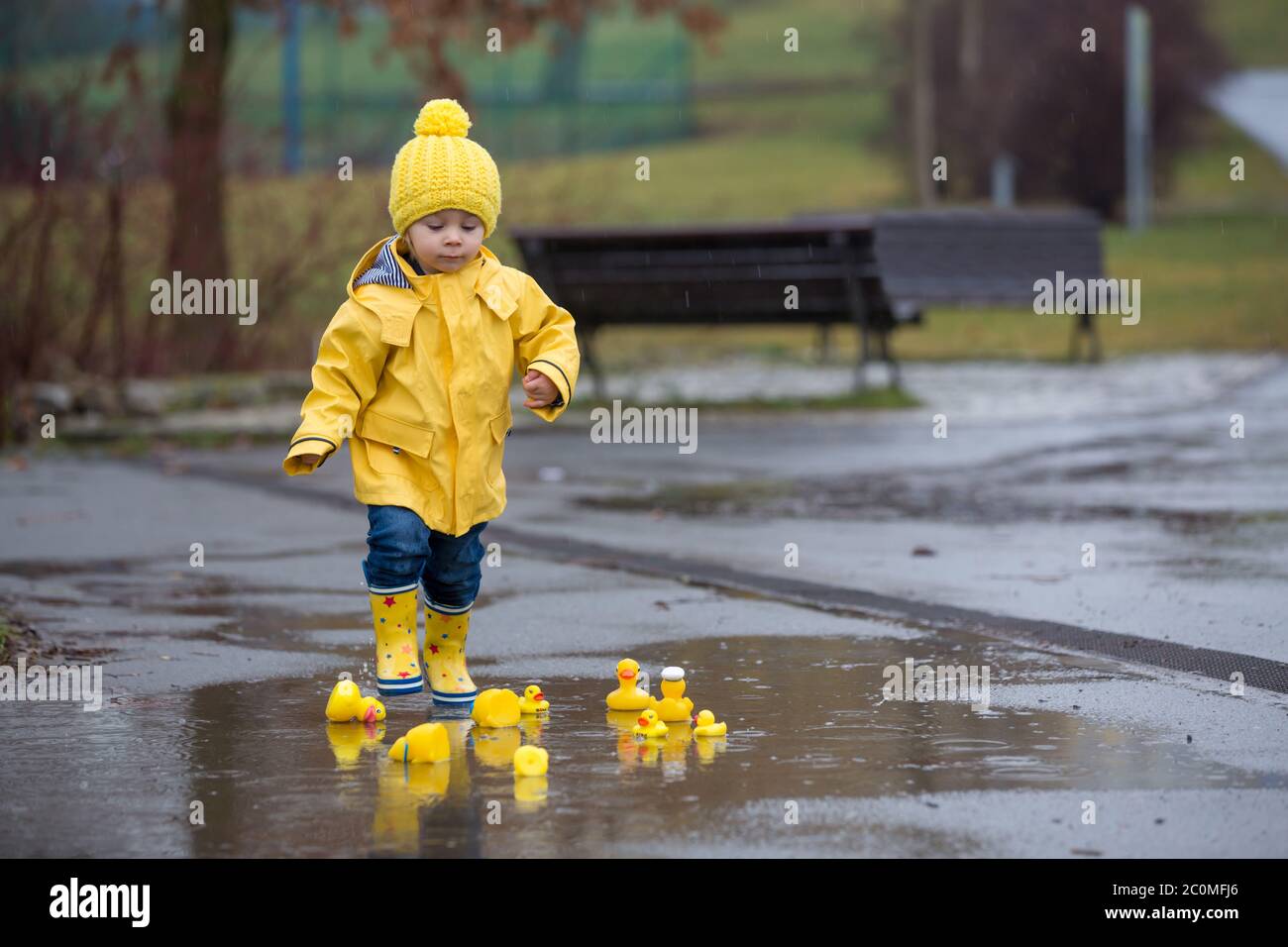 Beautiful funny blonde toddler boy with rubber ducks and colorful umbrella, jumping in puddles and playing in the rain, wintertime Stock Photo