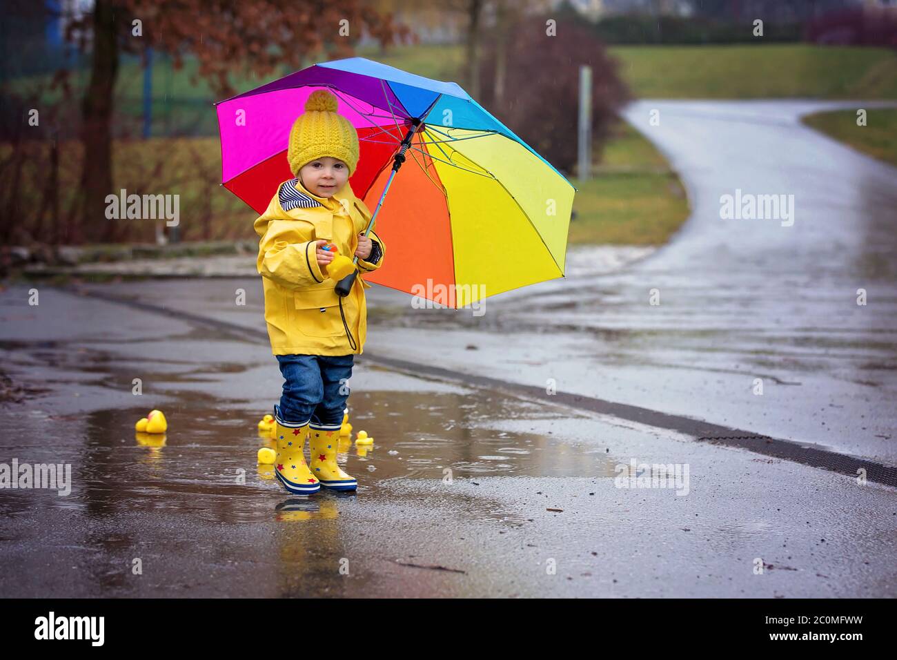 Beautiful funny blonde toddler boy with rubber ducks and colorful umbrella, jumping in puddles and playing in the rain, wintertime Stock Photo