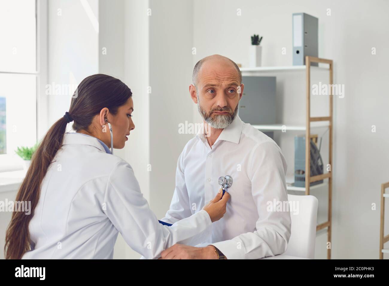 Woman doctor cardiologist listens with stethoscope to patient senor man in clinic office. Visit to the doctor in a medical hospital. Stock Photo