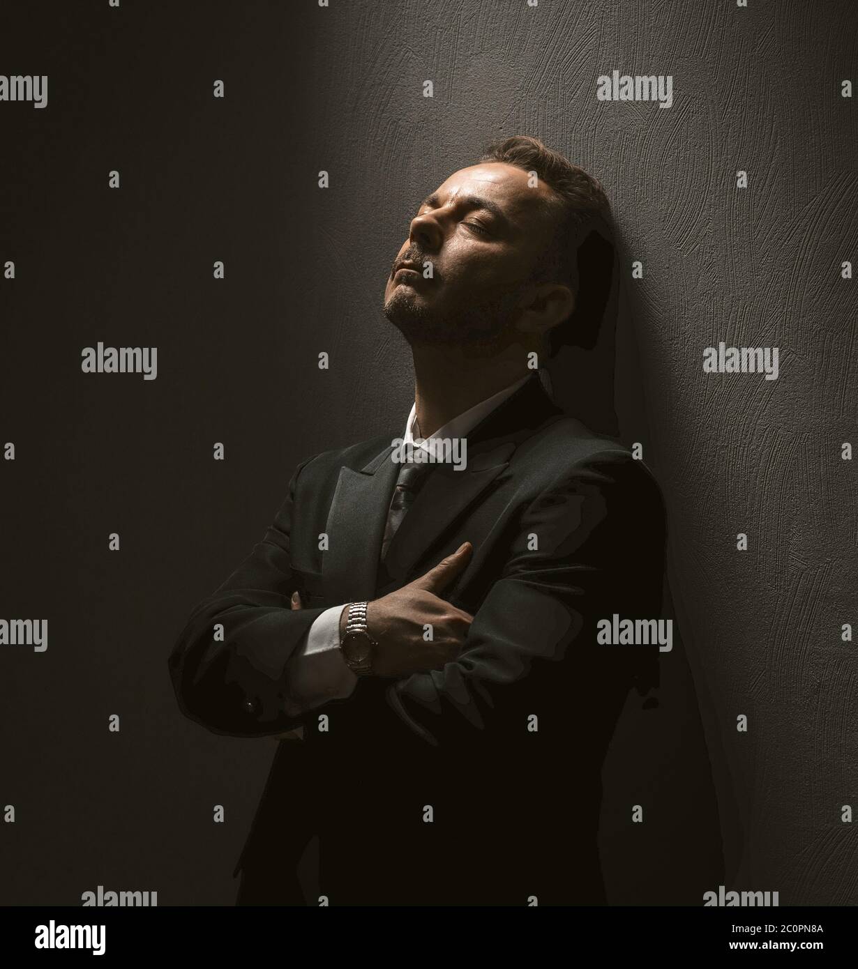 Thoughtful businessmans rests alone in dark room. Caucasian Man in dark suit with closed eyes stands arms crossed leaning against the wall. Stress Stock Photo