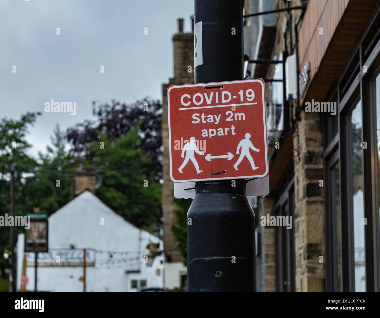 A red and white Covid-19 sign on a lamp post outside shops in Baildon, Yorkshire. The sign advises pedestrians to stay 2 metres apart. Stock Photo