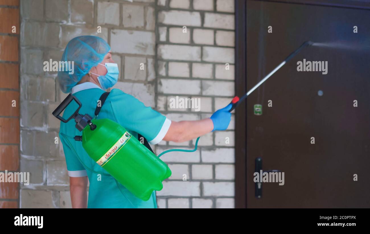 Masked woman cleaning door with liquid disinfectant using large bottle and spray. Antiseptic treatment concept. Preventive medical treatment. City Stock Photo