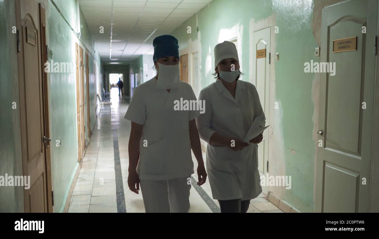 Two masked female nurses in white coats walking through hospital hall looking at camera. Hospital interior in process of repair. Abstract defocused Stock Photo