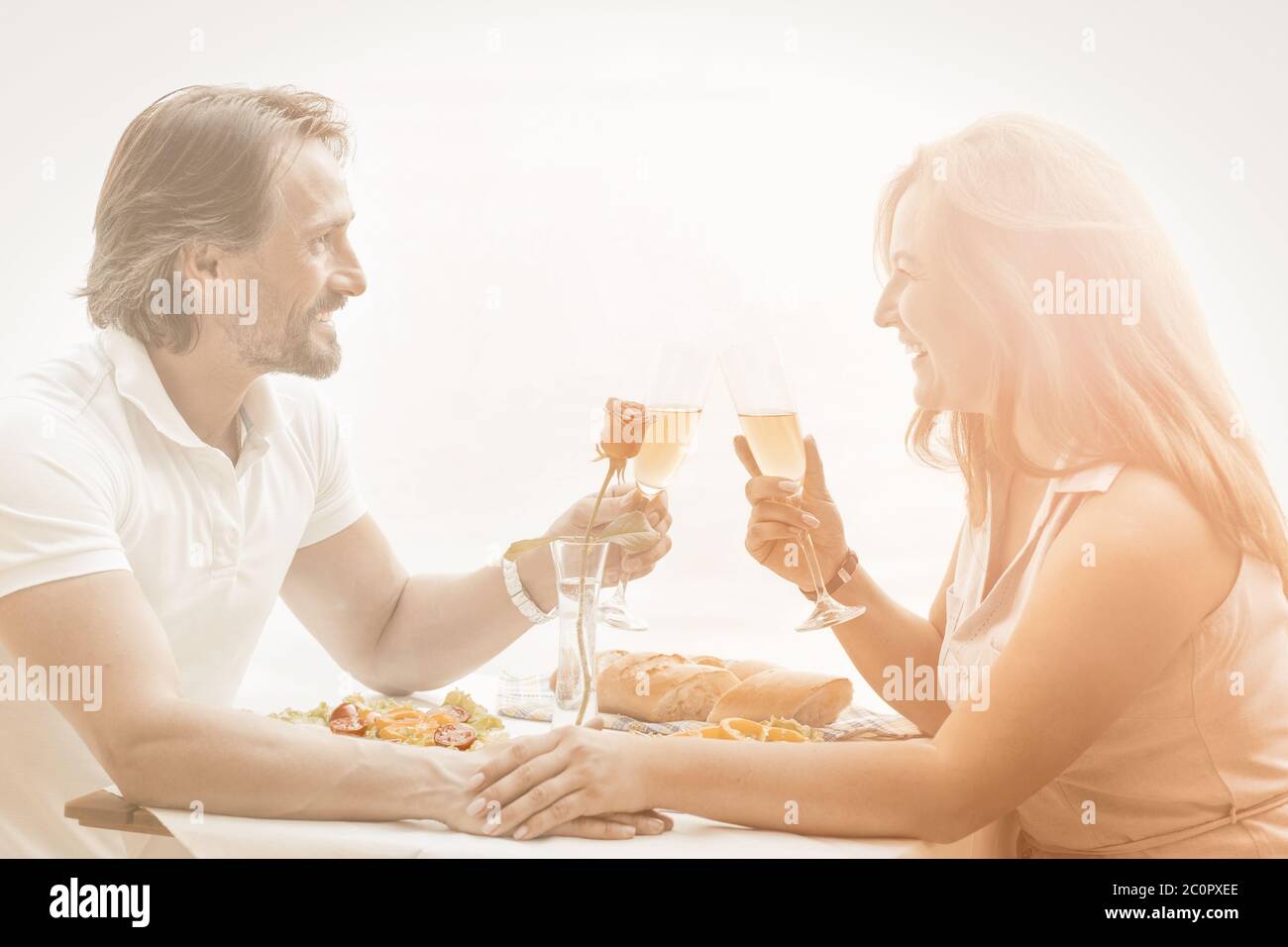 Man and woman holding glasses of white wine look at each other while sitting at table in fresh air. Happy couple dating at beach restaurant. Toned Stock Photo