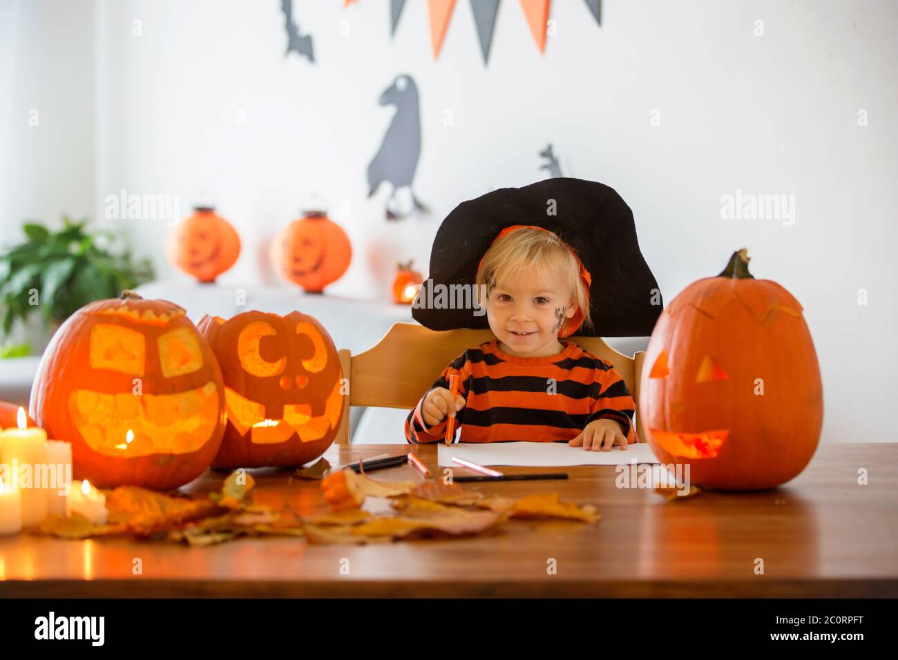 Child, toddler boy, drawing with pasteles pumpkin at home on Halloween, halloween carved pumpkin on the table, decoration on the wall Stock Photo