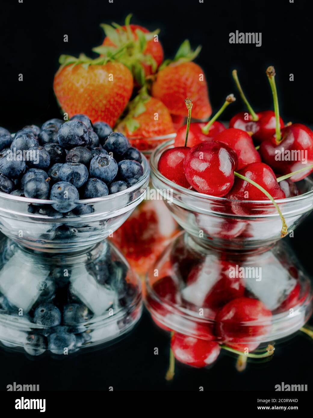 Three glass bowl filled with strawberries, cherries, and blueberries on a mirrow with reflection.  Black background. Stock Photo
