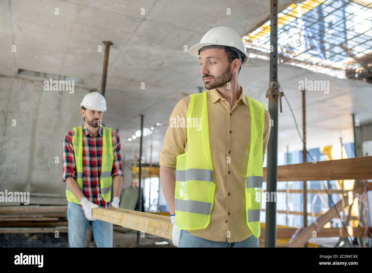Building workers in yellow vests and gloves carrying wooden board together Stock Photo