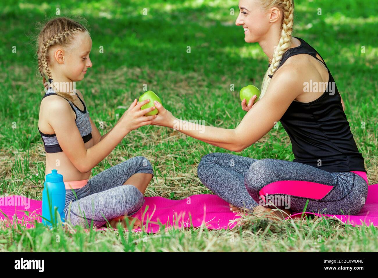 Mother is giving to daughter a green apple sitting on the roll mat in park. Stock Photo