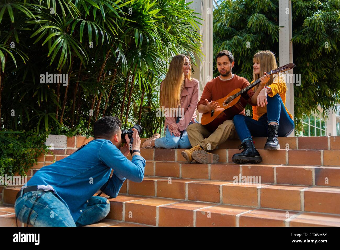 A musician with a guitar and two young girls pose on a staircase next to some trees for a photographer, space for text and natural light Stock Photo