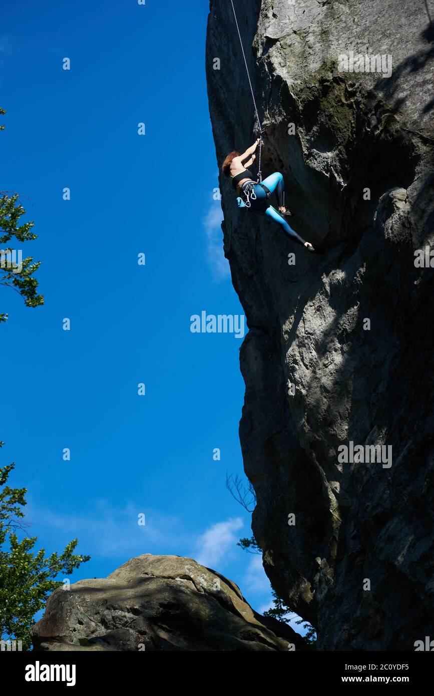 Vertical snapshot of female rock-climber, climbing up a cliff, wearing blue  leggings and a black top in safety harness, clear blue sky on background.  Low angle view. Extreme concept Stock Photo 