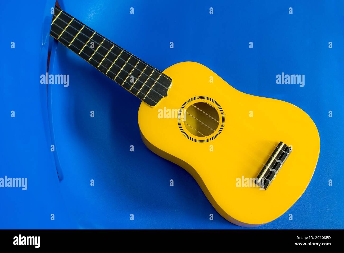 Yellow ukulele. Child uke on a blue plastic kids chair background. Musical instrument for children. Brightly colored ukulele for fun music play. Stock Photo